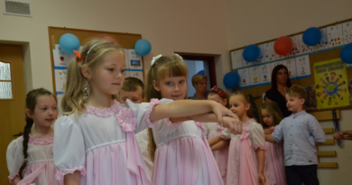 cztery dziewczynki w biało-różowych długich sukienkach i jeden chłopiec o ciemnych włosach w niebieskiej koszuli i czarnych spodniach. Dzieci ustawione są w rzędzie i trzymają się za ręce, tańcząc poloneza.