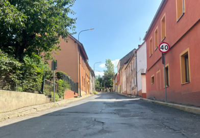 Ulica, z prawej strony bordowy budynek, z lewej krzewy, mur zaporowy oraz budynek w kolorze jasnego beżu.