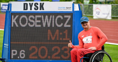 Niepełnosprawny mężczyzna w czerwonym dresie i niebieskiej czapce, siedzący na wózku inwalidzkim. Obok czarna tablica z wynikami .