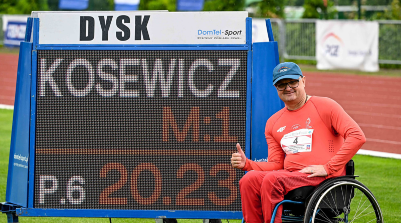 Niepełnosprawny mężczyzna w czerwonym dresie i niebieskiej czapce, siedzący na wózku inwalidzkim. Obok czarna tablica z wynikami .