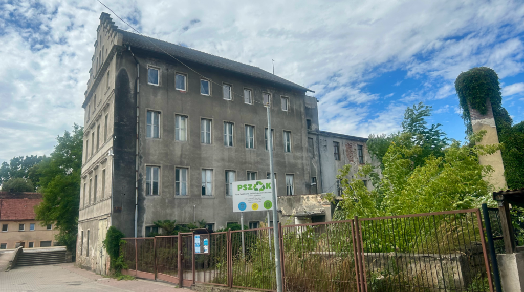 Strary budynek o 4 piętrach w kolorze szarym, koło obiektu tablica informacją z nazwą PSZOK oraz zielone krzewy