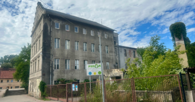 Strary budynek o 4 piętrach w kolorze szarym, koło obiektu tablica informacją z nazwą PSZOK oraz zielone krzewy