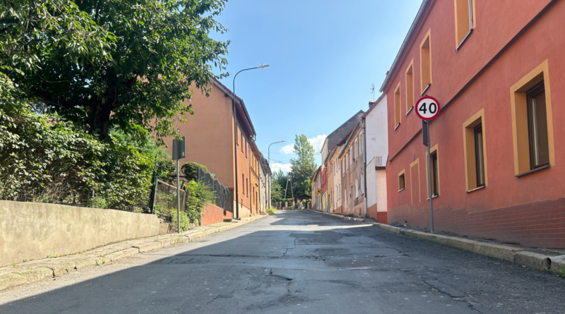 Ulica, z prawej strony bordowy budynek, z lewej krzewy, mur zaporowy oraz budynek w kolorze jasnego beżu.