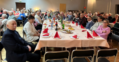 seniorzy siedzący przy stole, na nim czerwone serwetki, kwiaty, napoje oraz posiłki