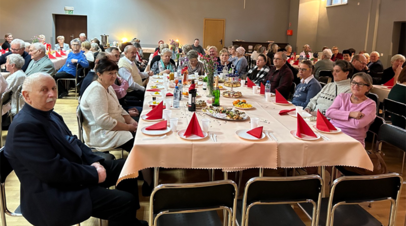 seniorzy siedzący przy stole, na nim czerwone serwetki, kwiaty, napoje oraz posiłki