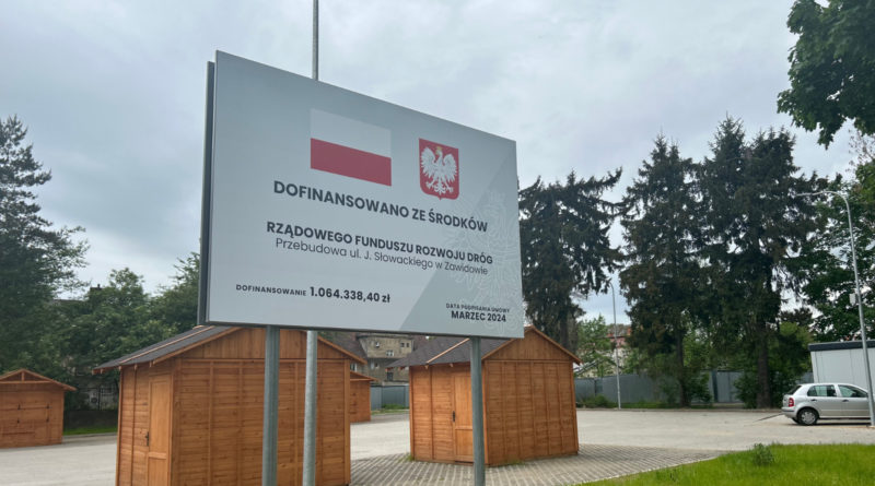tablica z informacją o dofinansowniu, stojąca przy placu targowym, za tablicą dwie drewniane budki handlowe. Na tablicy widoczne także flaga narodowa i godło Polski.