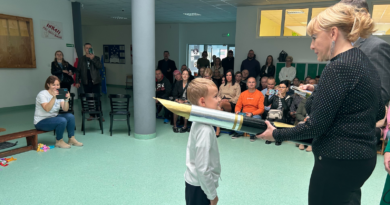 aula szkoły, na środku niej chłopiec w białej koszuli oraz kobieta ubrana na czarno, trzymająca duże symboliczne pióro. W tle rodzice i nauczyciele siedzący na ławkach.