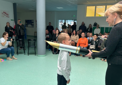 aula szkoły, na środku niej chłopiec w białej koszuli oraz kobieta ubrana na czarno, trzymająca duże symboliczne pióro. W tle rodzice i nauczyciele siedzący na ławkach.