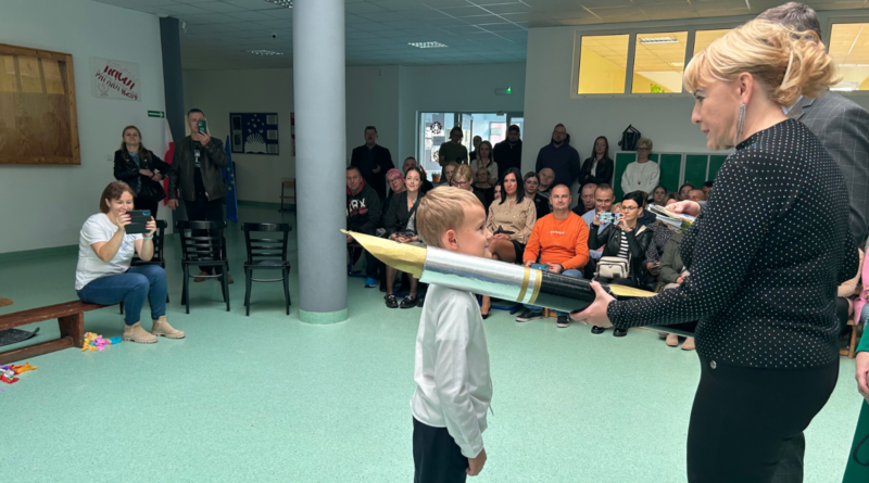 aula szkoły, na środku niej chłopiec w białej koszuli oraz kobieta ubrana na czarno, trzymająca duże symboliczne pióro. W tle rodzice i nauczyciele siedzący na ławkach.