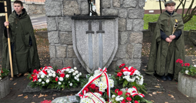 Obelisk z kamienia, na nim tarcza z dwoma mieczami. Pod pomnikiem biało-czerwone wiązanki kwiatów. Po prawej i lewej stronie pomnika chłopcy w zielonych mundurach z ramionami w dłoniach.