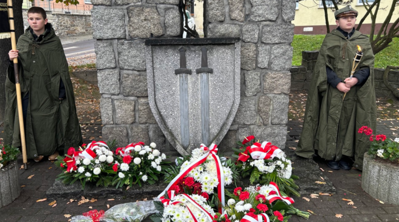 Obelisk z kamienia, na nim tarcza z dwoma mieczami. Pod pomnikiem biało-czerwone wiązanki kwiatów. Po prawej i lewej stronie pomnika chłopcy w zielonych mundurach z ramionami w dłoniach.