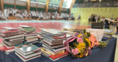 Nagrody książkowe na stole oraz bukiety kwiatów leżą na stole. W tle uczniowie na sali sportowej.