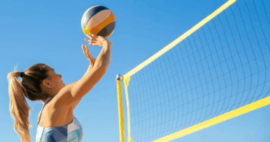 Kobieta w biało-niebieski bikini serwuje piłkę do gry w siatkówkę. Przed nią siatka do gry w piłkę plażową.