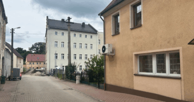 Dwa budynki, po prawej stronie pomarańczowy budynek mieszkalny, za nim 4-kontygnacyjny obiekt w kolorze szarym.