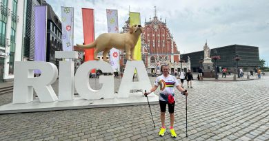 Mężczyzna w stroju spotowym i kijkami do uprawiania nordic walking, stojący na tle wielkiego napisu z nazwą miasta RIGA, w tle flagi i budynki.