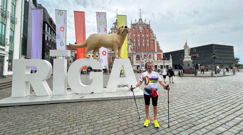 Mężczyzna w stroju spotowym i kijkami do uprawiania nordic walking, stojący na tle wielkiego napisu z nazwą miasta RIGA, w tle flagi i budynki.