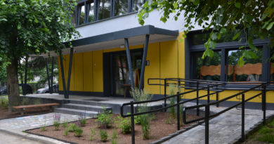 Żółty budynek przedszkola pośród drzew. Przed drzwiami frontowymi schody i barierki, obok po prawej stronie podjadę dla niepełnopsrawnych oraz pasaż zieleni.