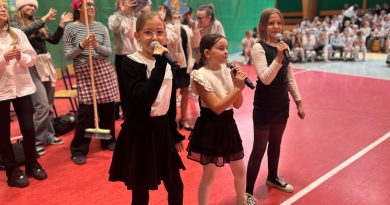 Trzy dziewczynki w stroju galowym śpiewające do mikrofonu. Z tyłu pozostałe dzieci.