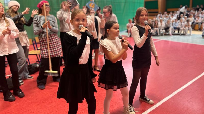 Trzy dziewczynki w stroju galowym śpiewające do mikrofonu. Z tyłu pozostałe dzieci.