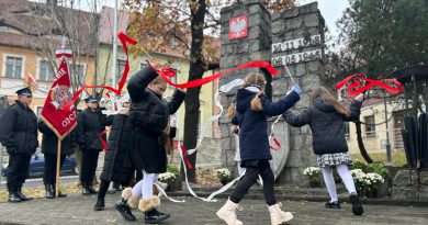 Dzieci tańczące z wstążkami w kolorach białym i czerwonym pod pod obeliskiem. W tle strażacy ze sztandarem.