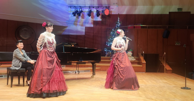 Dwie śpiewaczki operowe w czerwonych sukniach oraz mężczyzna przy fortepianie.