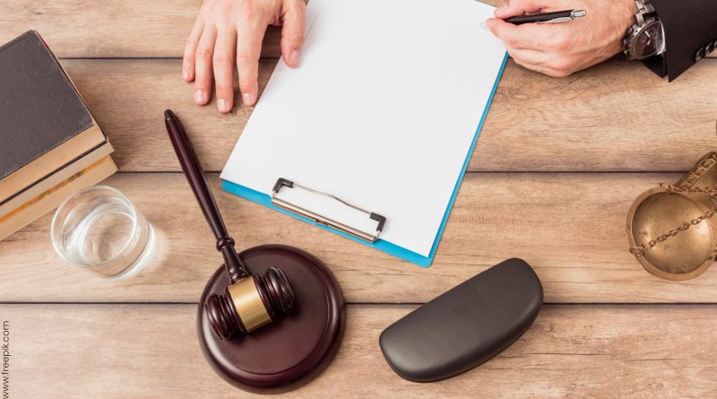 Tydzień Pomocy Ofiarom Przestępstw – dyżury w Prokuraturze Rejonowej w Zgorzelcu biurko a na nim biała kartka do notowania, męskie dłonie, młotek sędziowski, waga-symbol sprawiedliwości, etui na okulary, książka oraz szklanka z wodą.