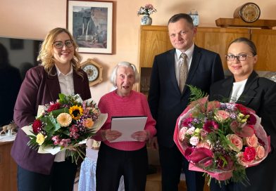Wyjątkowy jubileusz w Zawidowie – pani Honorata skończyła 100 lat! Trzy kobiety na zdjęciu oraz jeden mężczyzna. Dwie Panie trzymają bukiet kwiatów. Zdjęcie wykonane w pokoju.