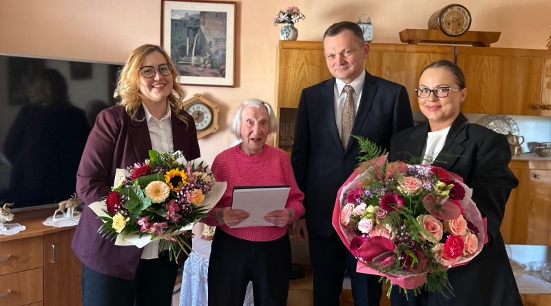 Wyjątkowy jubileusz w Zawidowie – pani Honorata skończyła 100 lat! Trzy kobiety na zdjęciu oraz jeden mężczyzna. Dwie Panie trzymają bukiet kwiatów. Zdjęcie wykonane w pokoju.