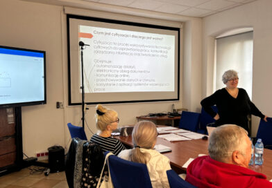 Z Internetem na Ty! Zawidowscy seniorzy wzięli udział w szkoleniu „e-Fajfy” sala konferencyjna, duży monitor, 3 osoby siedzący przy stołach i instruktorka uśmiechająca się do uczestników.