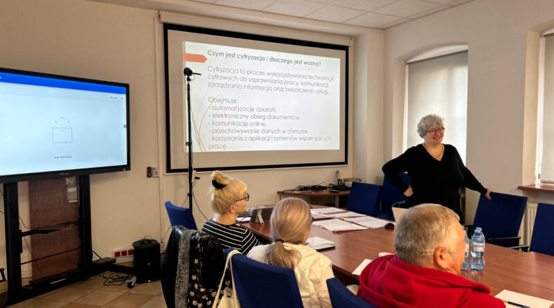Z Internetem na Ty! Zawidowscy seniorzy wzięli udział w szkoleniu „e-Fajfy” sala konferencyjna, duży monitor, 3 osoby siedzący przy stołach i instruktorka uśmiechająca się do uczestników.